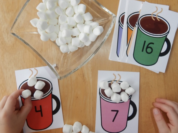 Two hands sort marshmallows onto illustrated cards with colored mugs and numbers. A glass bowl holds more marshmallows. Cards display numbers 4, 7, and 16. The activity is laid out on a wooden surface.