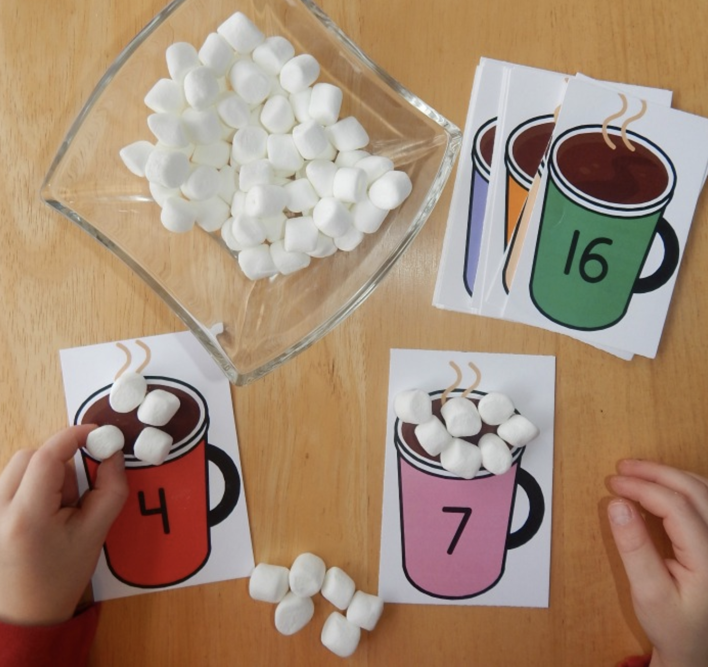 Two hands sort marshmallows onto illustrated cards with colored mugs and numbers. A glass bowl holds more marshmallows. Cards display numbers 4, 7, and 16. The activity is laid out on a wooden surface.