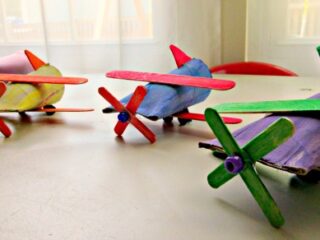 Colorful homemade toy airplanes made from recycled materials sit on a table. Each plane features a bright propeller, wings, and tail, painted in various colors. The background shows a softly lit room with curtains.