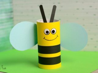 A handmade bee craft made from a yellow cardboard tube with black stripes, googly eyes, a smile, black antennae, and blue wings, placed on a green surface with a dotted background.