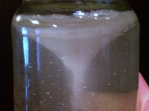 A glass jar filled with clear liquid showing a swirling whirlpool or vortex at the center. The background is dark, highlighting the movement within the jar.