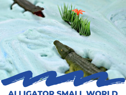 Two toy alligators on blue foamy surface with artificial grass and a small red flower. Text reads Alligator Small World Sensory Play with a website URL at the bottom.