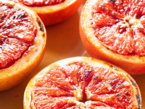 Halved grapefruits with caramelized tops are arranged in a white baking dish. The fruits vibrant red flesh glistens with a light glaze.