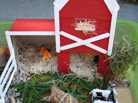A childrens farm playset featuring a red barn with a white roof, surrounded by toy animals including cows, a horse, and a rooster. The barn is decorated with hay and enclosed by a white fence, set on a base of pebbles and grass.