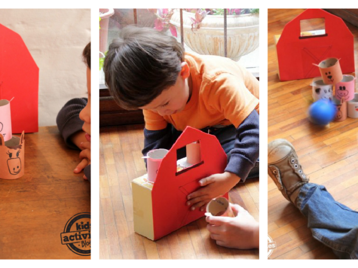 Children are playing a homemade farm animal themed bowling game. The setup uses a red barn made from cardboard and animal faces on paper rolls. A blue ball is rolled to knock them over on a wooden floor.