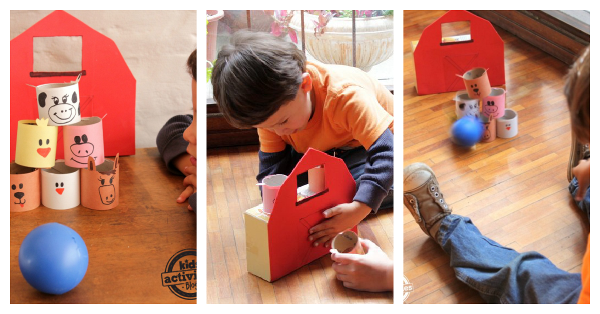 Children are playing a homemade farm animal themed bowling game. The setup uses a red barn made from cardboard and animal faces on paper rolls. A blue ball is rolled to knock them over on a wooden floor.