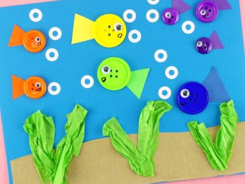A colorful underwater-themed craft made with buttons as fish, each with googly eyes and paper tails. Blue paper serves as the background, with white circles as bubbles and green tissue paper as seaweed on a sandy bottom.