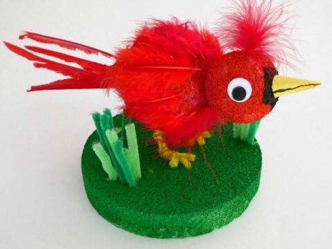 A handmade craft of a red cardinal bird using feathers, pipe cleaners, and a foam base. The bird features a yellow beak and stands on a green platform with decorative greenery. Text reads Cardinal Craft for Kids and craftulate.com.