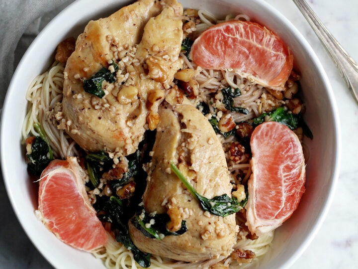 A bowl of angel hair pasta topped with chicken, spinach, walnuts, and grapefruit slices. A fork rests on the side of the bowl. Text reads: Chicken with Spinach and Grapefruit over Angel Hair along with a website URL at the bottom.
