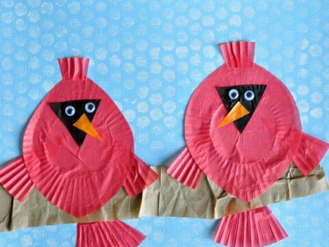A craft project featuring two red cardinals made from cupcake liners, with googly eyes and orange beaks, perched on a brown paper branch against a textured blue background.