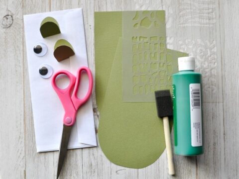 Craft supplies on a table: a pair of pink scissors, a foam brush, a green bottle of paint, googly eyes, green paper cutouts, and a stencil. Items are arranged on a white wooden surface.