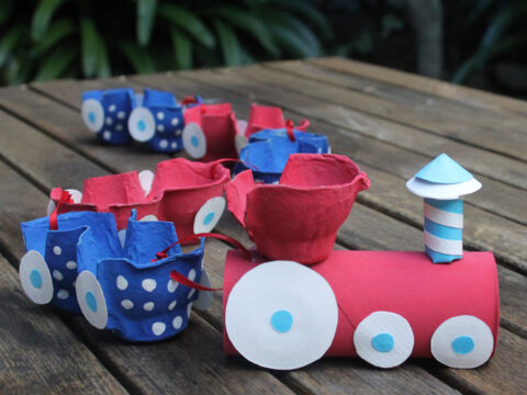 A colorful toy train made from painted egg cartons and a cardboard tube is on a wooden table. The train has bright red, blue, and white colors with white wheels, set against a blurred natural background.