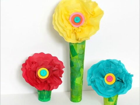 Three colorful paper flowers with button centers are displayed. The flowers are red, yellow, and blue, each secured in a green textured base made from paper rolls. They are crafted with layered petals, creating a vibrant, handmade decoration.