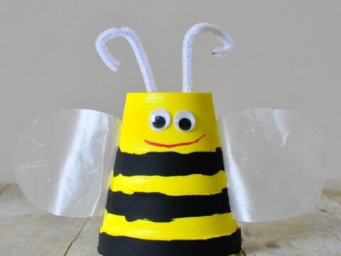 A cute bee craft made from a yellow and black striped cup with googly eyes, a red smile, white pipe cleaner antennae, and translucent wings. The background is a neutral shade.