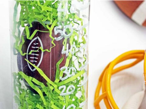 A clear bottle contains green shredded paper and a small toy football inside. The bottle is labeled with yard lines. Nearby are parts of a yellow helmet and a football. Text on the image reads Football Discovery Bottle.