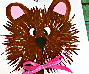 A cute bear face painted using a fork on a piece of paper, with pink inner ears and a pink polka-dot bow. The background is a teal wooden surface. Text on the image reads fork bear and sassydealz.com.