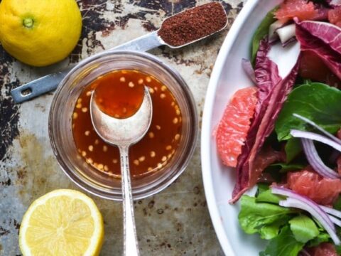 A top-down view of a salad with grapefruit slices, red onion, and leafy greens on a white plate. Beside it, a lemon, half a lemon, a measuring spoon with paprika, and a jar of dressing with a spoon.