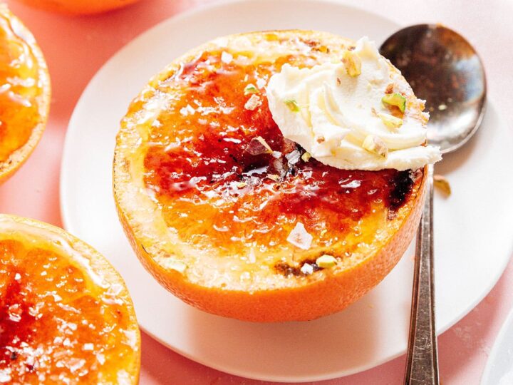 A halved grapefruit on a white plate, caramelized on top with a dollop of cream and sprinkled pistachios. A vintage spoon rests beside it. Other grapefruit halves and glasses in the background on a pink surface.