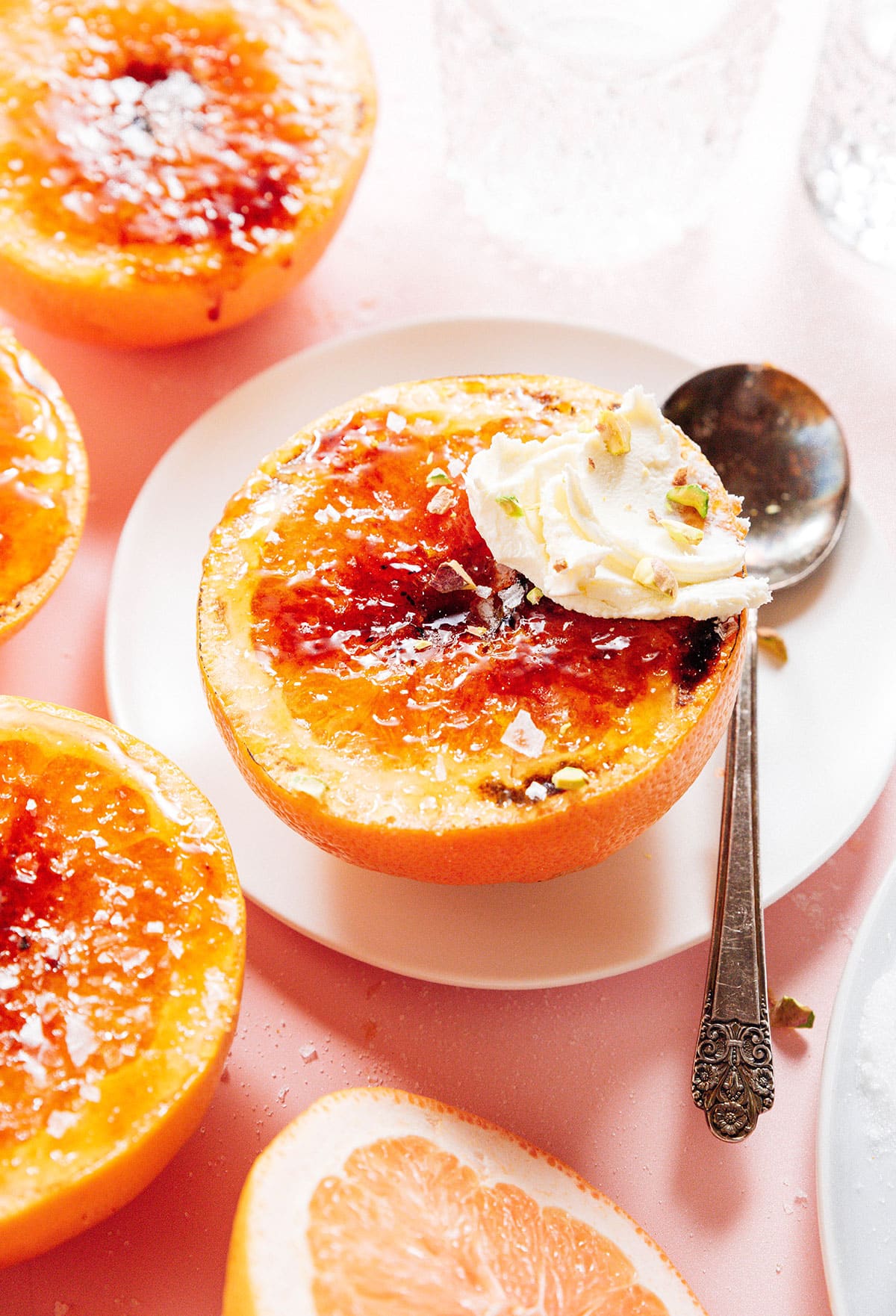 A halved grapefruit on a white plate, caramelized on top with a dollop of cream and sprinkled pistachios. A vintage spoon rests beside it. Other grapefruit halves and glasses in the background on a pink surface.