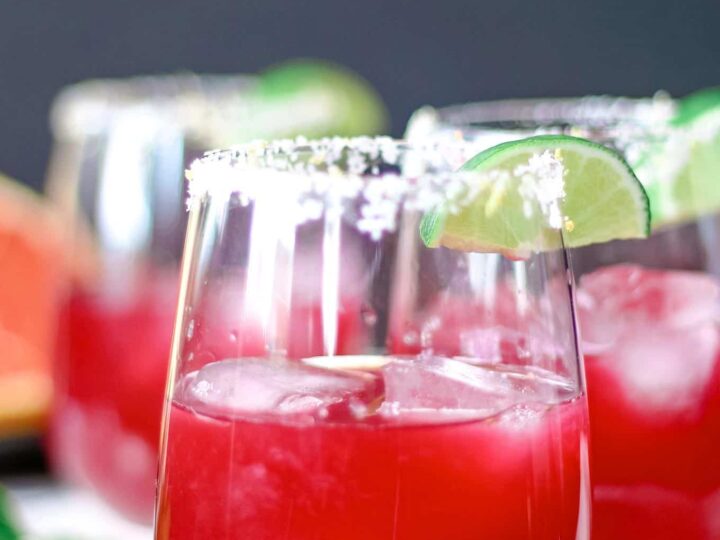 Two glasses filled with a bright red cocktail, garnished with lime wedges and a salted rim, sit on a white surface. The drinks contain ice cubes, and the background is softly blurred.