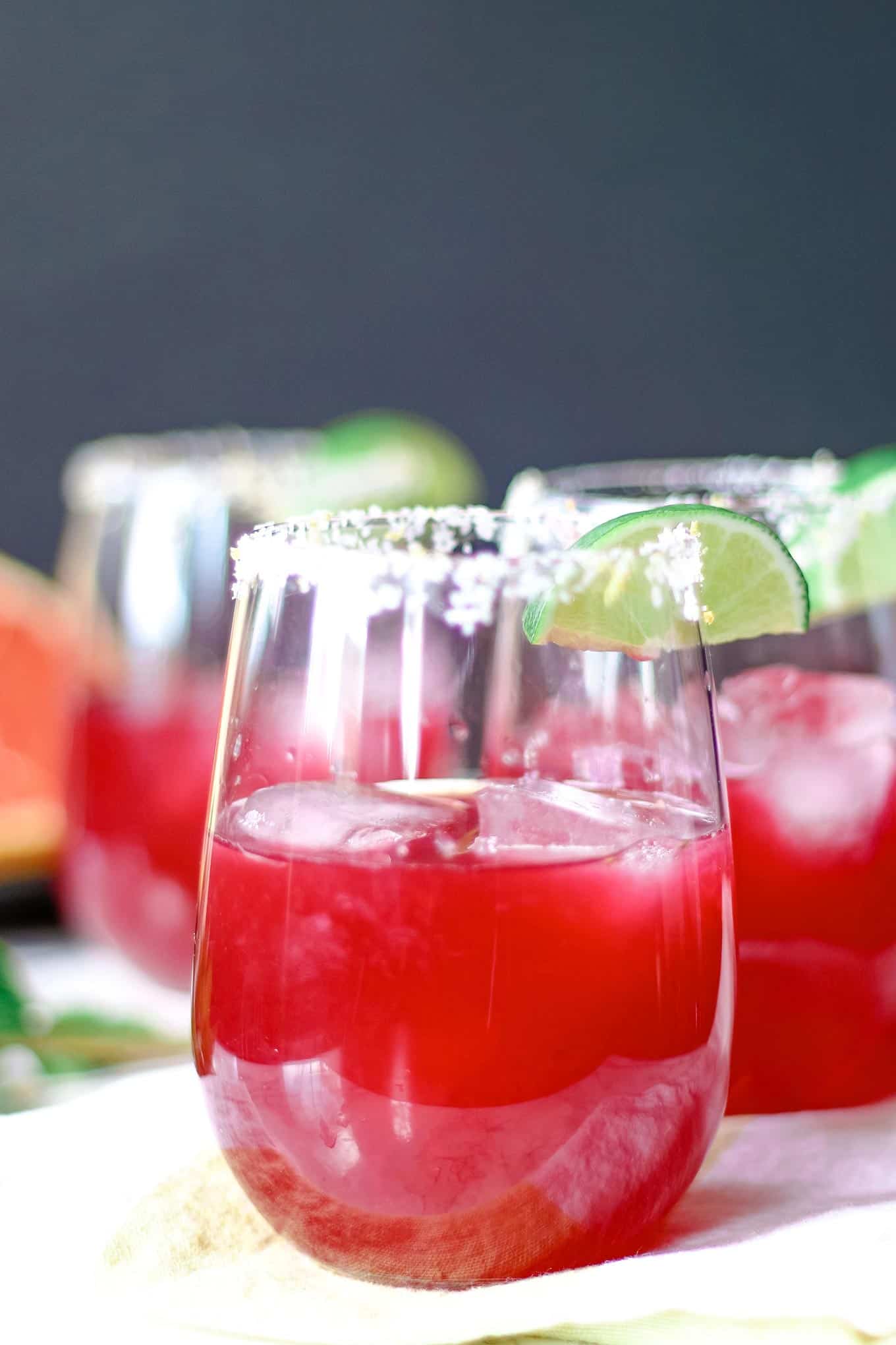 Two glasses filled with a bright red cocktail, garnished with lime wedges and a salted rim, sit on a white surface. The drinks contain ice cubes, and the background is softly blurred.