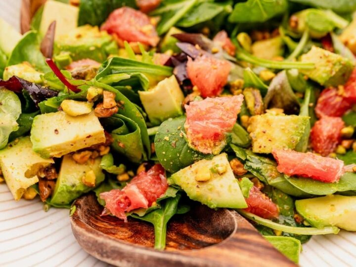 A vibrant salad with spinach, avocado, grapefruit segments, and pistachios, served on a white plate with a wooden serving spoon.