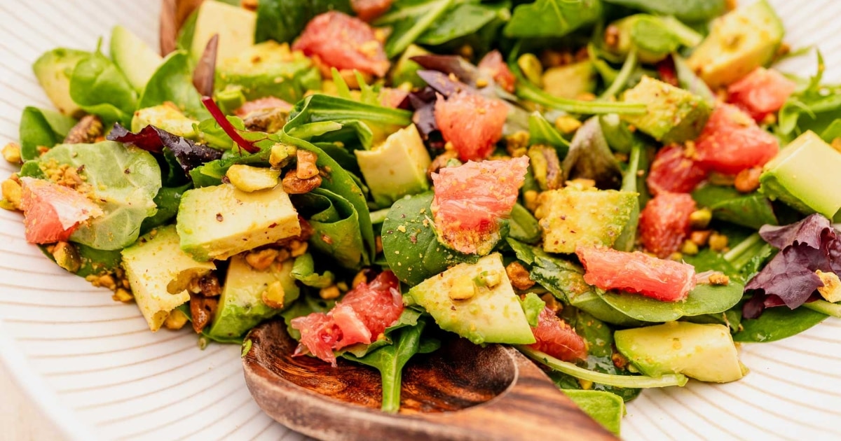 A vibrant salad with spinach, avocado, grapefruit segments, and pistachios, served on a white plate with a wooden serving spoon.