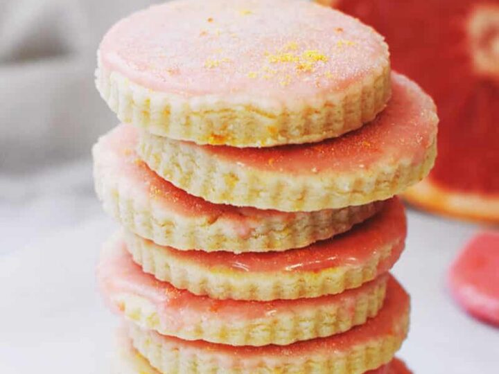 A stack of grapefruit-flavored cookies with pink icing and scalloped edges is arranged on a light surface. A sliced grapefruit is partially visible in the background.
