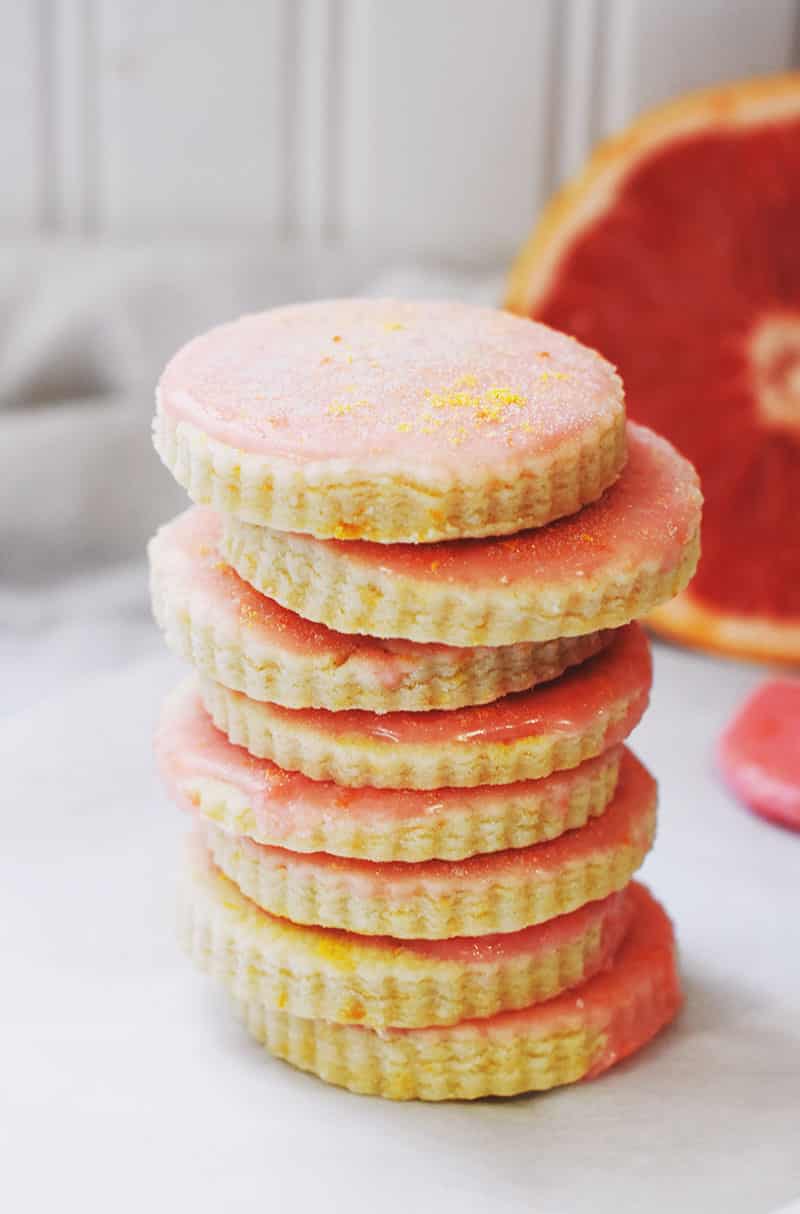 A stack of grapefruit-flavored cookies with pink icing and scalloped edges is arranged on a light surface. A sliced grapefruit is partially visible in the background.