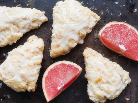 Five triangular glaze-covered scones arranged on a dark surface with two sections of a sliced red grapefruit.