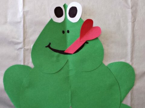 A paper frog craft made from green heart shapes, with large googly eyes and a red tongue sticking out. Text at the top reads Heart Frog Craft. Website watermark at the bottom.
