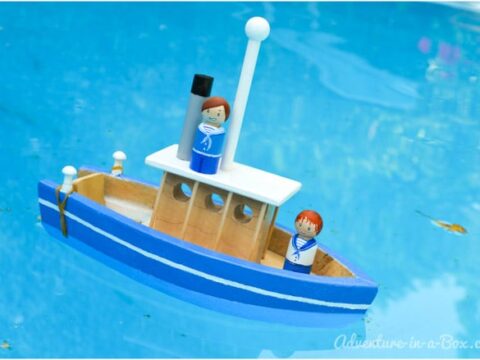 A small wooden toy boat floating in a pool. It is painted blue and white with two peg people figures on board, each wearing blue outfits. The water is clear and has a few small leaves floating on the surface.