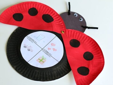 A ladybug craft made from red and black paper plates with black spots. The wings reveal a white circular paper divided into four sections with drawings of a mailbox, bubbles, hearts, and a gift. Two black pipe cleaners serve as antennae.