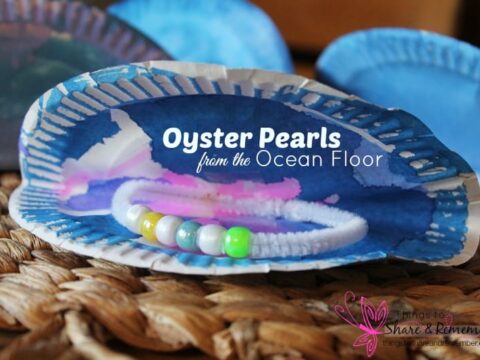 A paper plate craft resembling an oyster with painted blue and white hues. Inside, a bracelet made of colorful beads is placed, mimicking pearls. Text on the oyster reads, Oyster Pearls from the Ocean Floor. A woven surface is underneath.