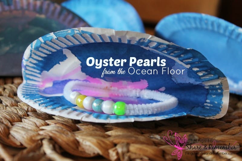 A paper plate craft resembling an oyster with painted blue and white hues. Inside, a bracelet made of colorful beads is placed, mimicking pearls. Text on the oyster reads, Oyster Pearls from the Ocean Floor. A woven surface is underneath.