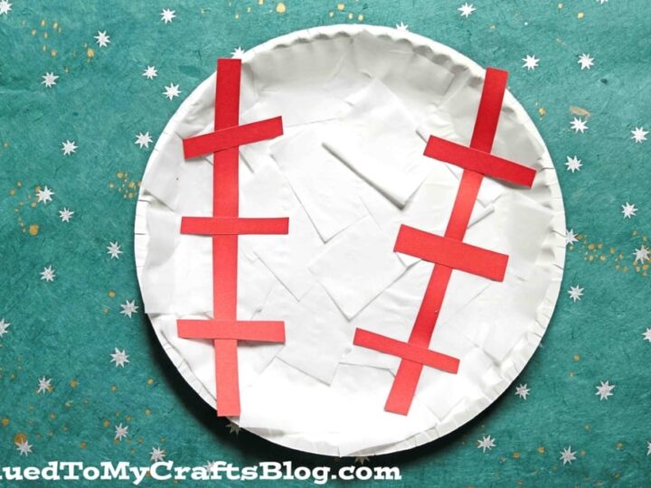 A paper plate crafted to look like a baseball, with white tissue paper covering the surface and red paper strips forming stitches. The background is a green surface with white star-like patterns. The text GluedToMyCraftsBlog.com is at the bottom.