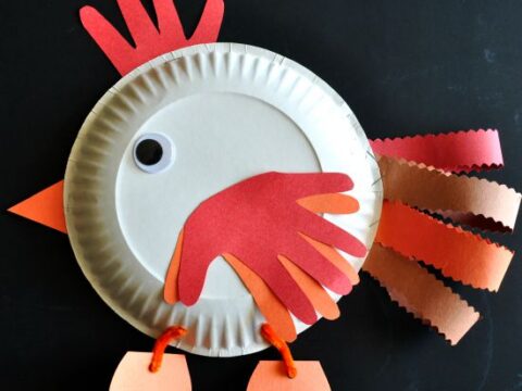 A paper plate craft turkey with colorful paper feathers and wings, a googly eye, and orange legs. Tail feathers are made from red, orange, and brown paper strips.