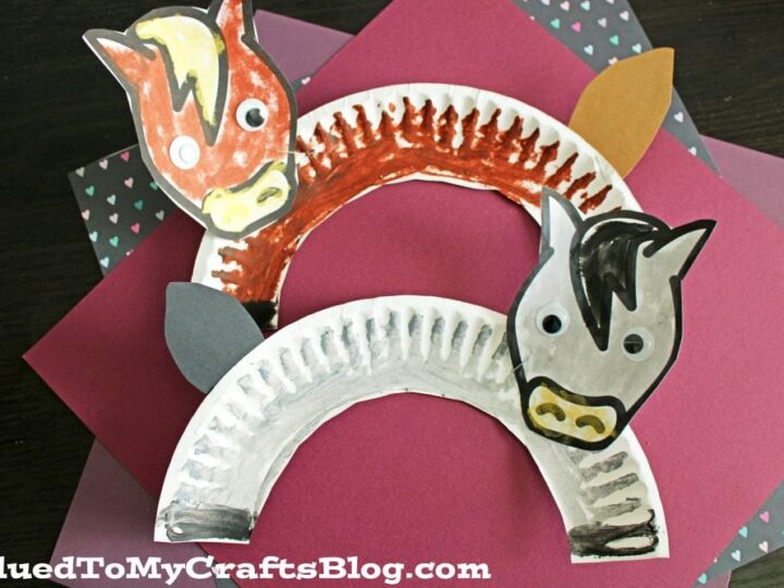 Two paper plate horse crafts with hand-painted faces and manes, one red and one grey, are arranged on dark and pink card stock backgrounds. Each horse has googly eyes and colored ears. The bottom background has a website watermark.