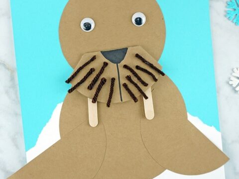 A craft project of a cute walrus made from brown paper and yarn, with googly eyes and popsicle sticks for tusks, set against a blue and white background.