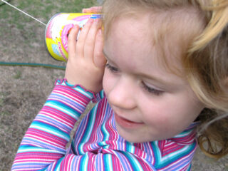 A young girl with curly hair is holding a tin can to her ear, playing with a homemade string telephone. She is outdoors, wearing a colorful striped long-sleeve shirt, smiling, and listening intently.