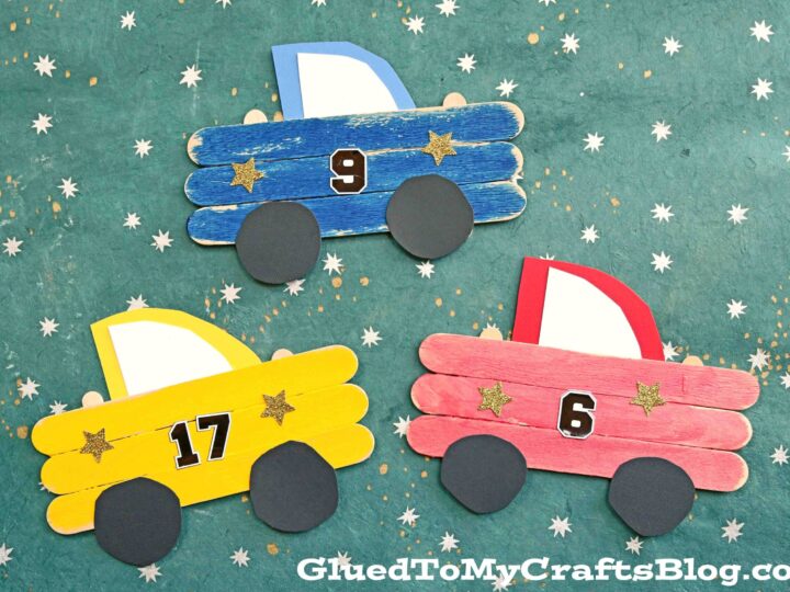 Three colorful popsicle stick race cars with numbers 9, 17, and 6. The cars are blue, yellow, and red, featuring black wheels. The background is a green floor with small star patterns.