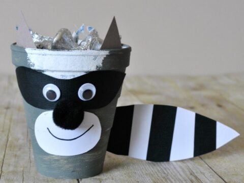 A raccoon-themed craft made from a gray flower pot featuring a painted mask, googly eyes, a paper nose, and a smile. A striped tail is attached to the pots side. iheartcraftythings.com is written at the bottom.