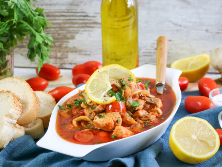 A dish of seafood stew garnished with lemon slices is presented in a white bowl. Surrounding it are slices of bread, fresh tomatoes, a bottle of olive oil, a bunch of parsley, lemon halves, and garlic on a blue cloth.