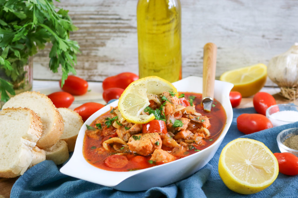 A dish of seafood stew garnished with lemon slices is presented in a white bowl. Surrounding it are slices of bread, fresh tomatoes, a bottle of olive oil, a bunch of parsley, lemon halves, and garlic on a blue cloth.