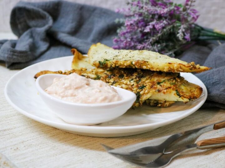 A white plate with crispy herb-crusted bread pieces and a white bowl of creamy dip. A folded gray napkin and purple flowers serve as background decor. Silverware is placed beside the plate on a beige tablecloth.