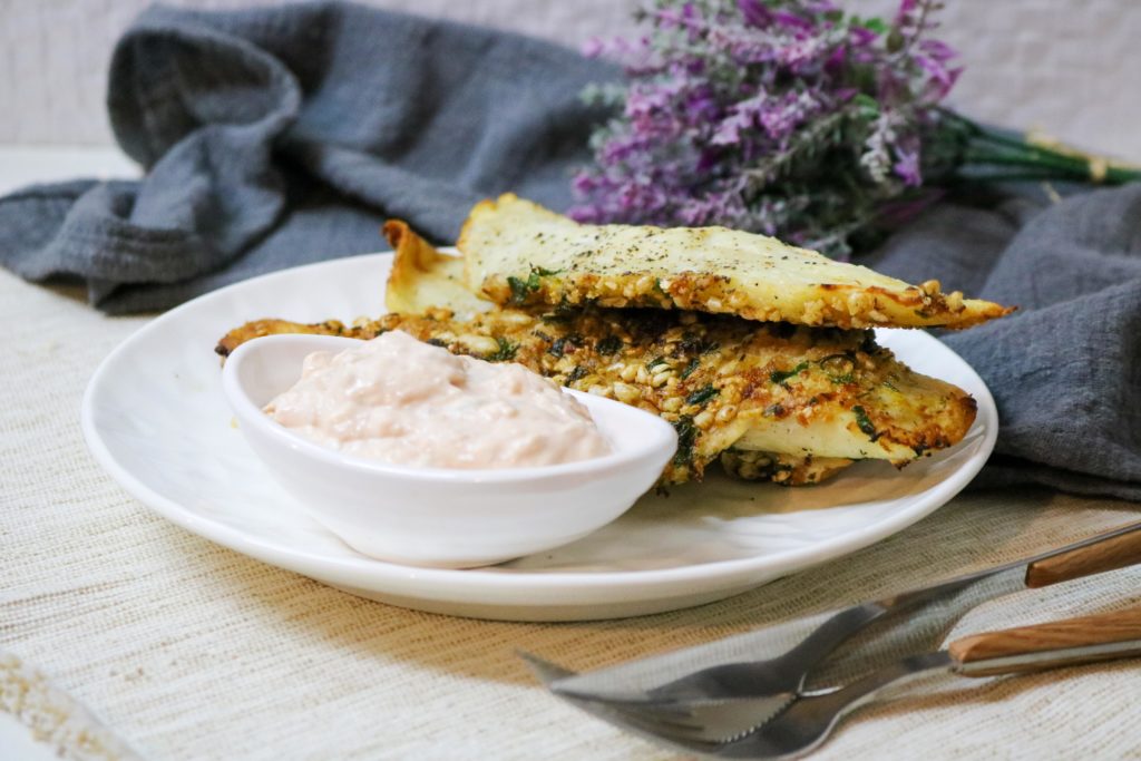 A white plate with crispy herb-crusted bread pieces and a white bowl of creamy dip. A folded gray napkin and purple flowers serve as background decor. Silverware is placed beside the plate on a beige tablecloth.