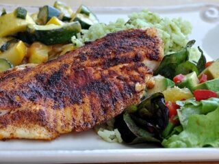 A seasoned and grilled fish fillet served on a plate with a side of mixed sautéed vegetables, green rice, and a fresh salad with lettuce, cucumbers, and tomatoes.