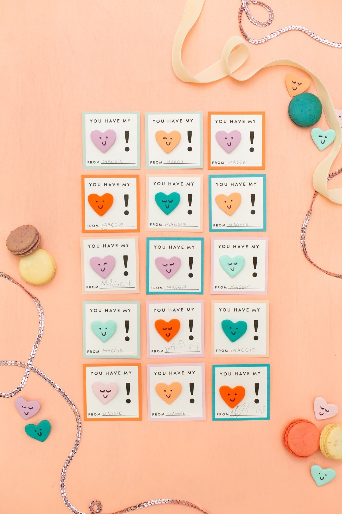 A grid of colorful cards on a peach background, each with a heart and the message You have my heart! Different colors and heart shapes. Surrounded by macarons, small heart trinkets, ribbon, and silver beads.