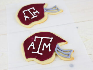 Two cookies shaped like football helmets with maroon icing and ATM written in white. They have gray icing details resembling helmet faceguards. The cookies are on a white paper on a light wooden surface.
