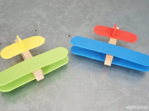 Two small, handmade toy airplanes made from colored popsicle sticks and clothespins on a gray textured surface. One plane is red and blue, and the other is yellow and green.